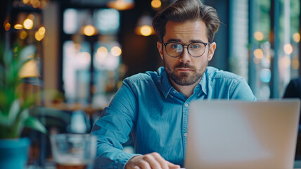 Young businessman works at his desk on a laptop, dressed in a blue shirt, with glasses and a notepad beside him, looking at the screen. - obrazy, fototapety, plakaty