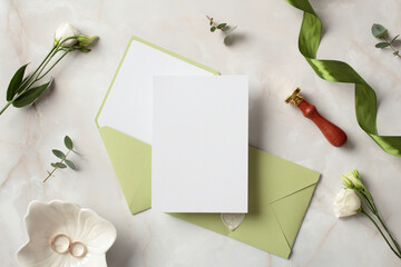 Wedding stationery set. Pastel green envelopes, blank paper card, gold rings, ribbon, wax stamp, flowers on stone background. Flat lay, top view, copy space