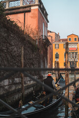 Gondolier stands in gondola, ready to glide onto Grand Canal. Gondola surrounded by old wooden poles with a vine-covered wall to the left. Yellow Venetian palazzo across the canal.