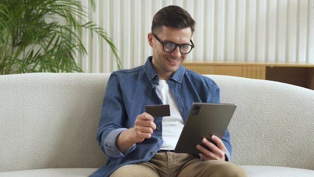 A man uses a bank card to pay for his purchases online and a tablet to use at home, on the couch.
