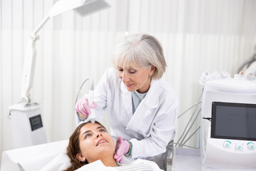 Careful old woman doctor conducting vacuum cleaning of young woman patient's face in aesthetic...