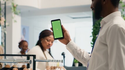 African american man holding mockup cellphone in premium clothing store with stylish formalwear...