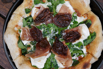 Top view of an arugula pizza with burrata cheese, sun dried tomatoes and cured raw ham, served in the pizza pan on the wooden table.