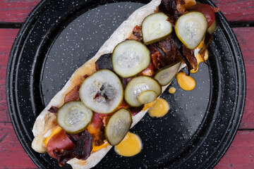 American food. Fusion. Closeup view of a gourmet hot dog with crispy bacon, cheddar cheese and cucumber slices, presented on a black dish on the wooden table.	
