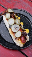 American food. Fusion. Closeup view of a gourmet hot dog with crispy bacon, cheddar cheese and cucumber slices, presente don a black dish on the red wooden table.