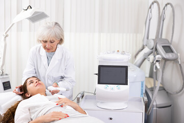 Careful aged woman doctor conducting laser therapy of young woman patient's face in aesthetic clinic