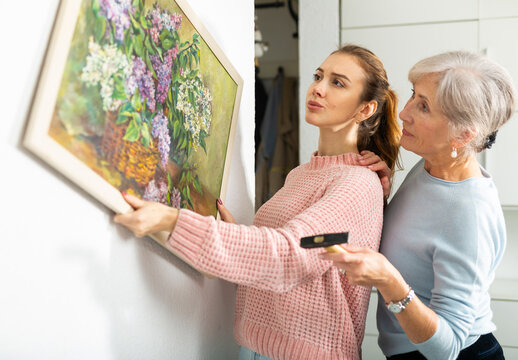 Young girl and older woman hanging together painting with depicted bouquet of lilacs on wall indoors .