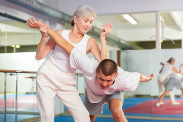 Focused elderly woman performing elbow strike and wristlock, painful control move to immobilize male opponent during self defense training in gym - 763621050