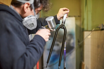 Latin young man wearing a respirator mask concentrated spray painting a bike fork in his workshop, applying a protective clear coat to give a professional finish to the process.
