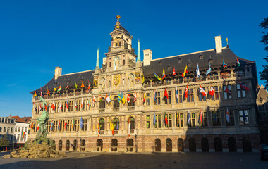 View of Brabo fountain and four-story Renaissance building of Antwerp City Hall with main facade lavishly decorated with sculptures and waving flags located on central Grote Markt square, Belgium..