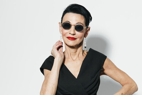 Elegant senior woman in sunglasses and black dress posing in front of white wall for camera portrait