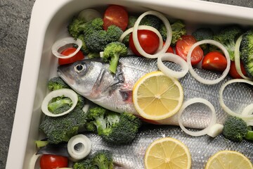 Raw fish with vegetables and lemon in baking dish on grey table, top view