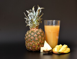 A mixture of fruit juices in a tall faceted glass on a black background, next to pieces of ripe pineapple and mango.