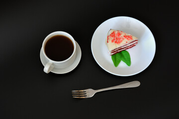 A plate with a piece of Red velvet cake, a fork and a cup of hot black coffee on a black background.