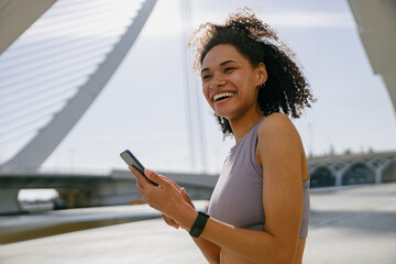 Smiling woman athlete in sportswear standing outdoors after training and use mobile phone