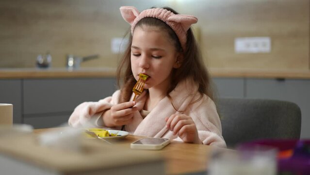 Slow motion. A girl, in a bathrobe and a pink cosmetic hair band with cat ears, sits at the table and eats pancake with filling. The girl looks at the screen of a mobile phone that lies on the table