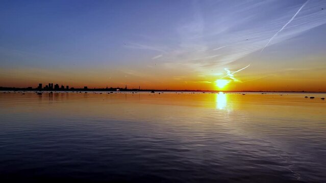 Tallin in sunset, setting over the water, creating a beautiful and serene atmosphere. The sky is filled with clouds and birds, water is calm and still. The scene is peaceful and calming