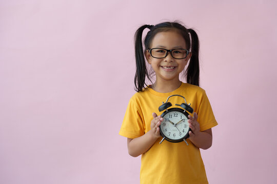 Image of a cheerful smiling little girl standing alone on a pink background holding an alarm clock. Time management concepts, learning, management.