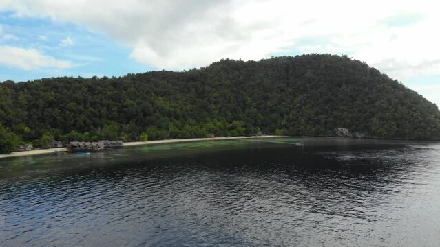 Hilly island landscape, overgrowth vegetation over sandy beach with wooden guesthouses. Aerial pan over Lumba Lumba guesthouses on Kri Island in Raja Ampat.
