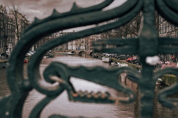 moody photo of a view of the Amsterdam canal seen through an iron railing with a cruise boat in the...
