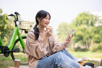 A happy young Asian woman is chilling in a green park, eating a donut and using her smartphone.