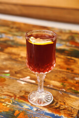 Crystal Glass with Refreshing Beverage and Lemon Slice. A clear, cut crystal glass filled with a deep amber colored liquid and garnished with a slice of lemon, resting on a textured, multicolored wood