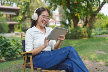 Fototapeta premium The concept of relaxation, lifestyle, and holiday, at the park. natural places Young Asian women wearing headphones and watching a movie on a tablet having fun, and relaxing.