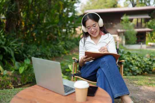 Young women wearing glasses wearing headphones reading a book Making diary notes Video calls via laptop online, relaxing, lifestyle, vacation, at the park Happily natural place.