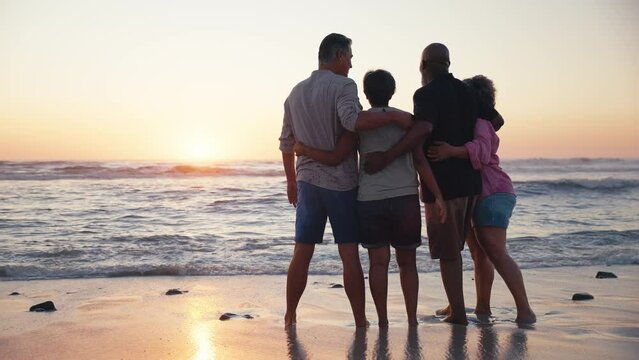 Rear view of mature couple with friends standing on beach shoreline looking out to sea at sunset - shot in slow motion