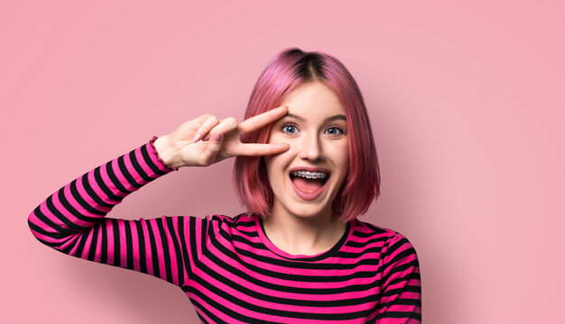 Photo image - happy beautiful woman in braces show v-sign victory peace hand gesture two fingers, with open mouth, isolated against pink wall background. Student girl at studio. Dental care ad concept