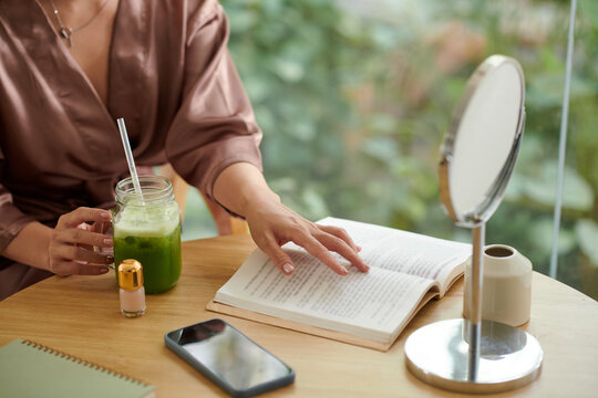 Cropped image of woman in silk robe reading novel and drinking green smoothie