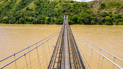 Obraz premium Western Bridge in Santa Fe de Antioquia, Colombia, spanning a muddy river with lush green hills in the background