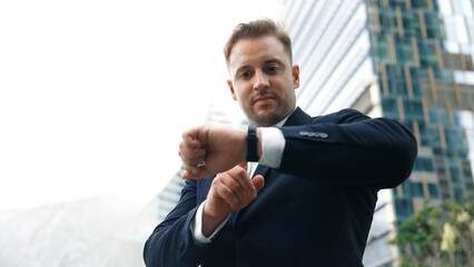 Low angle camera of businessman looking at watch to remind about time. Smart caucasian businessman checking time for being on time in important appointment or meeting with modern skyscraper. Urbane.