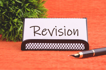 Revision word on a white business card on an orange background, next to a fountain pen and a plant...