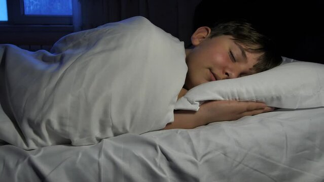 A cute eleven-year-old boy sleeping in a bed with white pillows charmingly folds his palms on the pillow and dreams. Good healthy sleep for the child. Comfortable bed and mattress with pillows