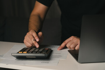 Asian man is using a calculator to calculate his family's monthly miscellaneous expenses at home and record them in his laptop computer.