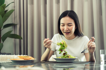 Asian young woman smiling as she scoops a salad on a plate and eats happily at home.