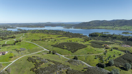 Drone image over beautiful green rolling hills in Northern California with New Hogan Lake in the background and a blue sky. - obrazy, fototapety, plakaty