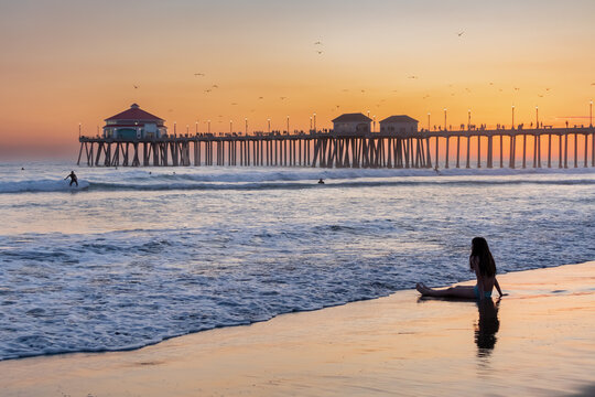 Silhouette of Teenage Girl Sitting in Wet Sand Watching Distant Surfers in Pacific Ocean at Twilight, March 2024, Huntington Beach, California, USA, horizontal