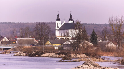 Charming European Village by the River in Spring, Tranquil Scene with White Church and Blue Sky