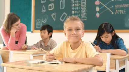 Caucasian smart child smiling at camera while doing classwork at classroom while happy multicultural student doing test or writing note in classroom at elementary class. Education concept. Pedagogy.