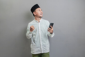 Asian Muslim man clenching his fist and holding his smartphone while looking at an empty space