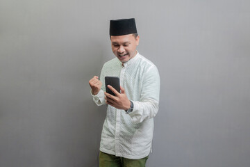 Asian Muslim man clenching his fist and holding his smartphone while looking to smartphone