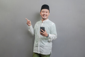 Asian Muslim man smilling while ponting to empty space while holding smartphone