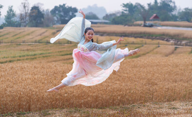 Flying dancer in the air. Happy Asian girl ballerina in a Chinese costume making a big jump in a golden barley field in the evening