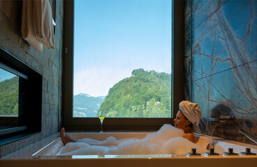 Woman Lying in a Bathtub with a Glass of Champagne and Enjoy the Window View over Mountain in a Sunny Summer Day in Burgenstock, Nidwalden, Switzerland.