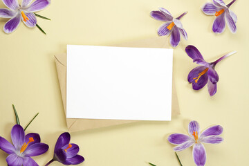Mockup invitation, blank paper greeting card, envelope and purple crocus flowers on a beige background. Flower background. Flat lay, top view. Space for text. 