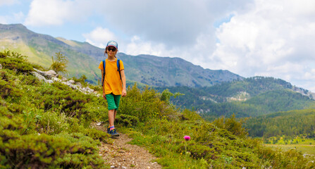 hiking in the mountains. A little boy with a backpack walks along a path against a background of mountains and clouds. Active healthy lifestyle on weekend hike journey.