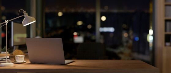 A laptop in a contemporary office room at night illuminated by dim lights from a table lamp.