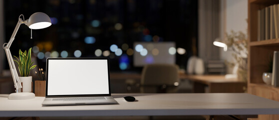 An office desk features a laptop computer mockup in a modern skyscraper office at night.